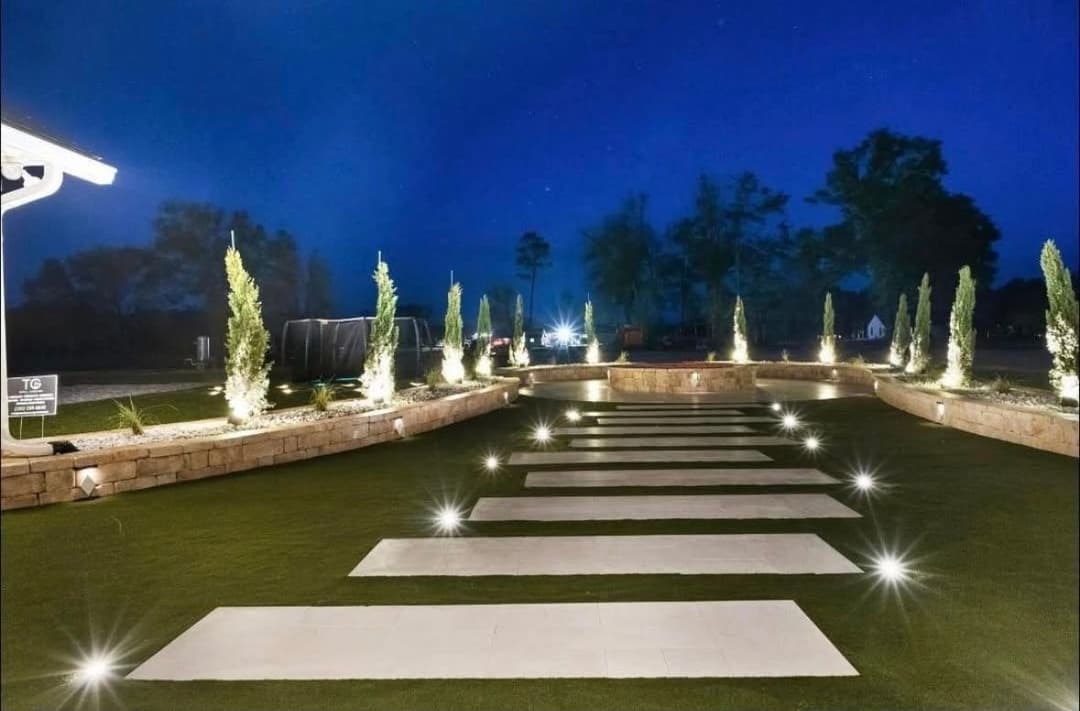 Illuminated stone pathway in landscaped garden at night, surrounded by greenery and lights.
