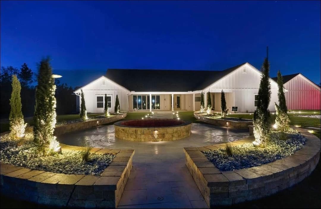 Modern countryside home at night with illuminated landscaping and circular stone feature.