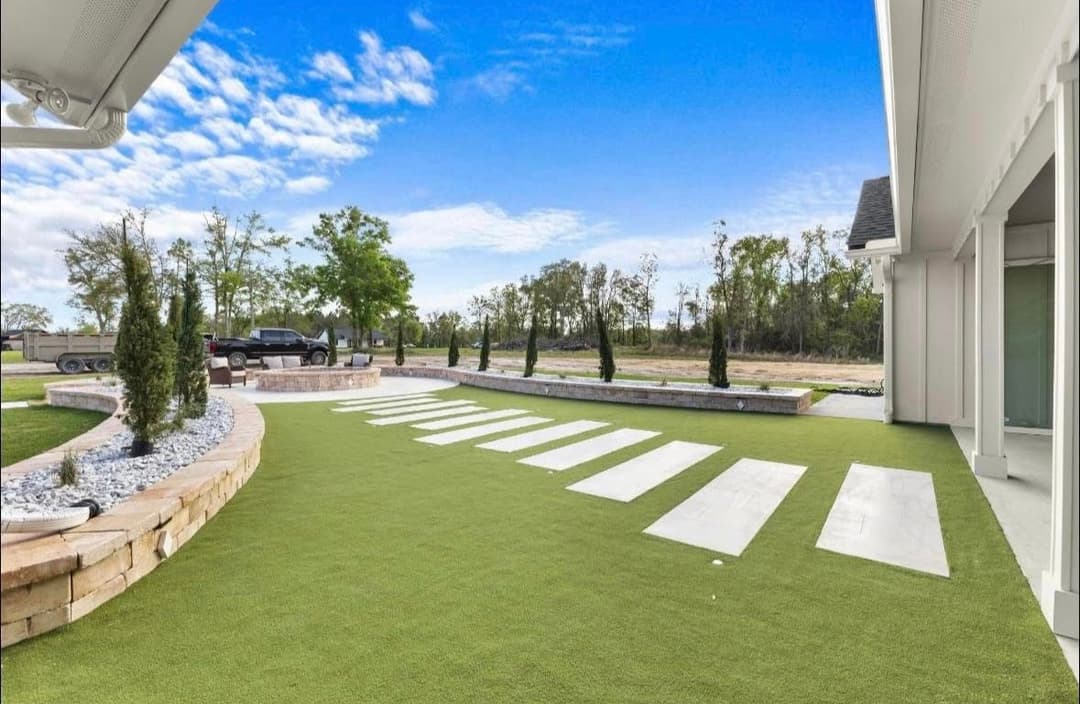 Modern outdoor landscape with green turf, stone pathways, and trees under a blue sky.