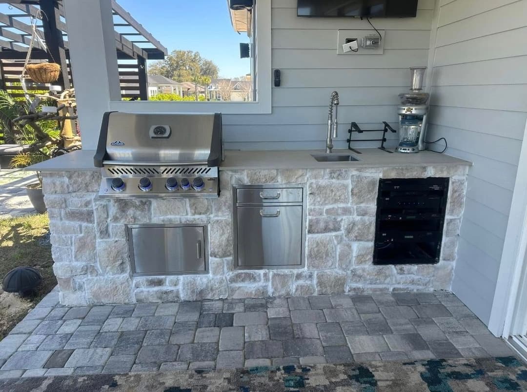 Outdoor kitchen with stainless steel grill, sink, and beverage cooler against a stone backdrop.