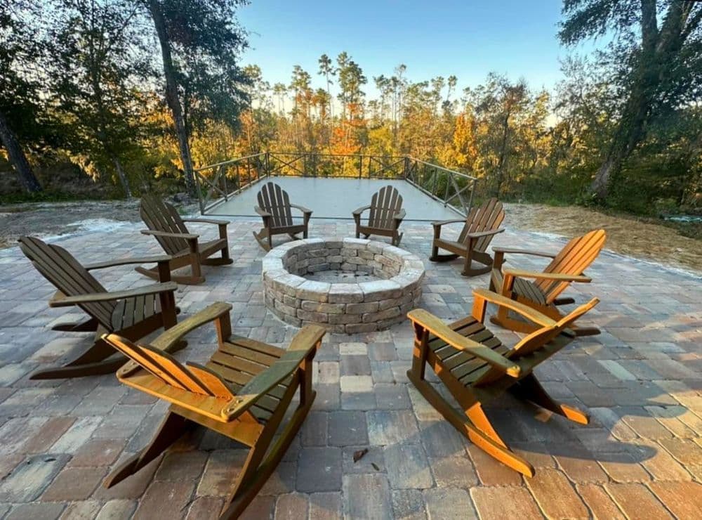 Adirondack chairs arranged around a stone fire pit in a wooded outdoor setting.