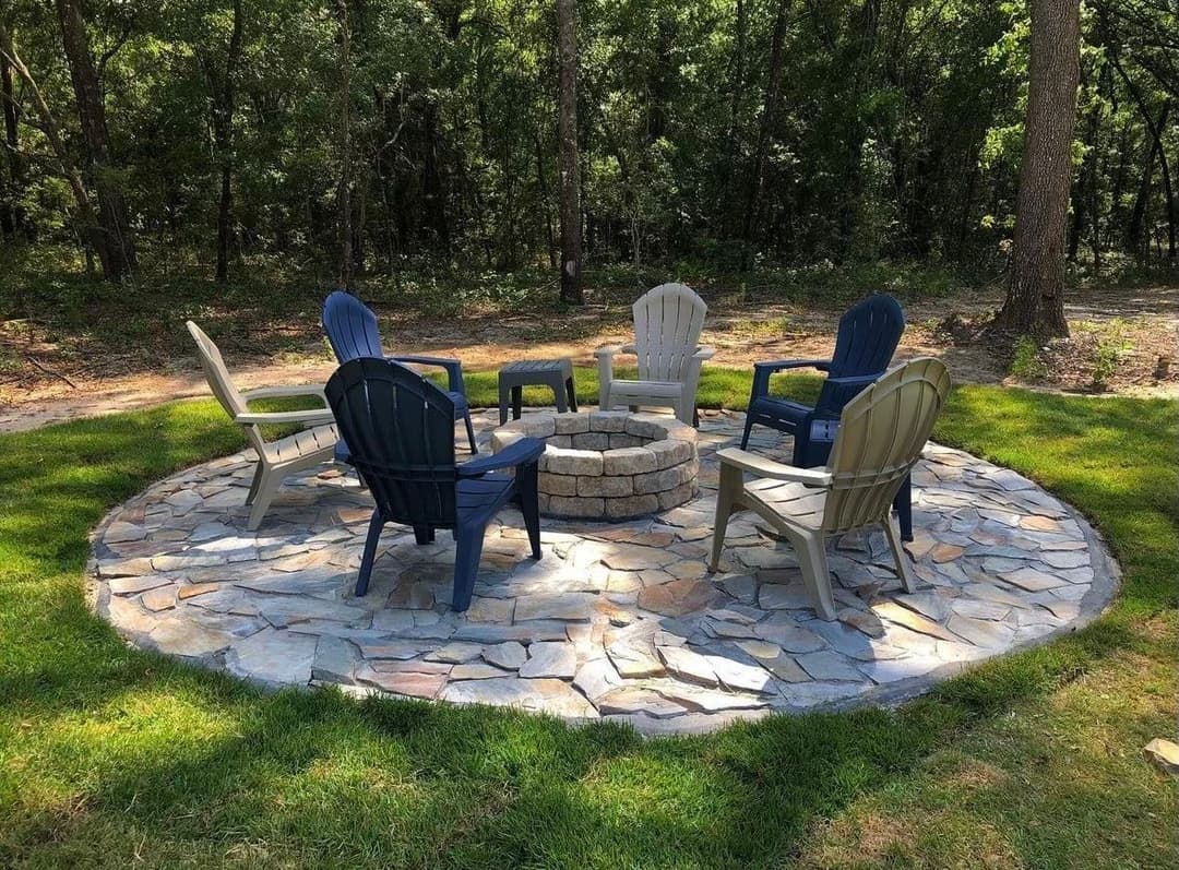 Outdoor fire pit seating area with stone circle and various chairs in a wooded setting.