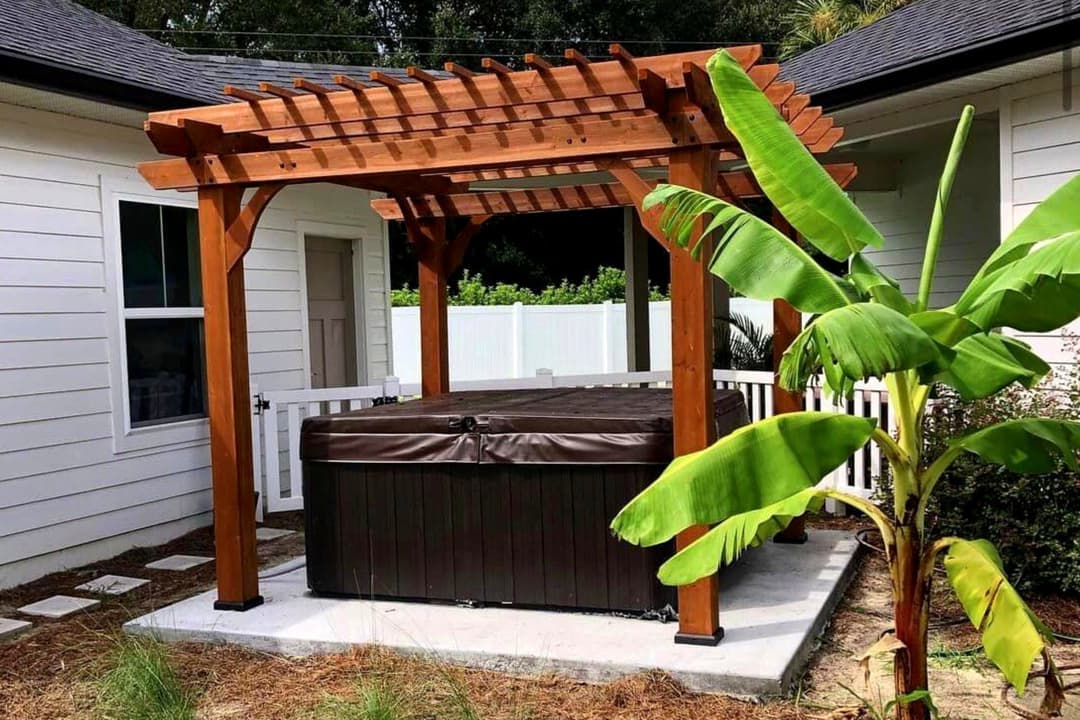 Wooden pergola over a hot tub surrounded by greenery and a banana plant in a backyard setting.
