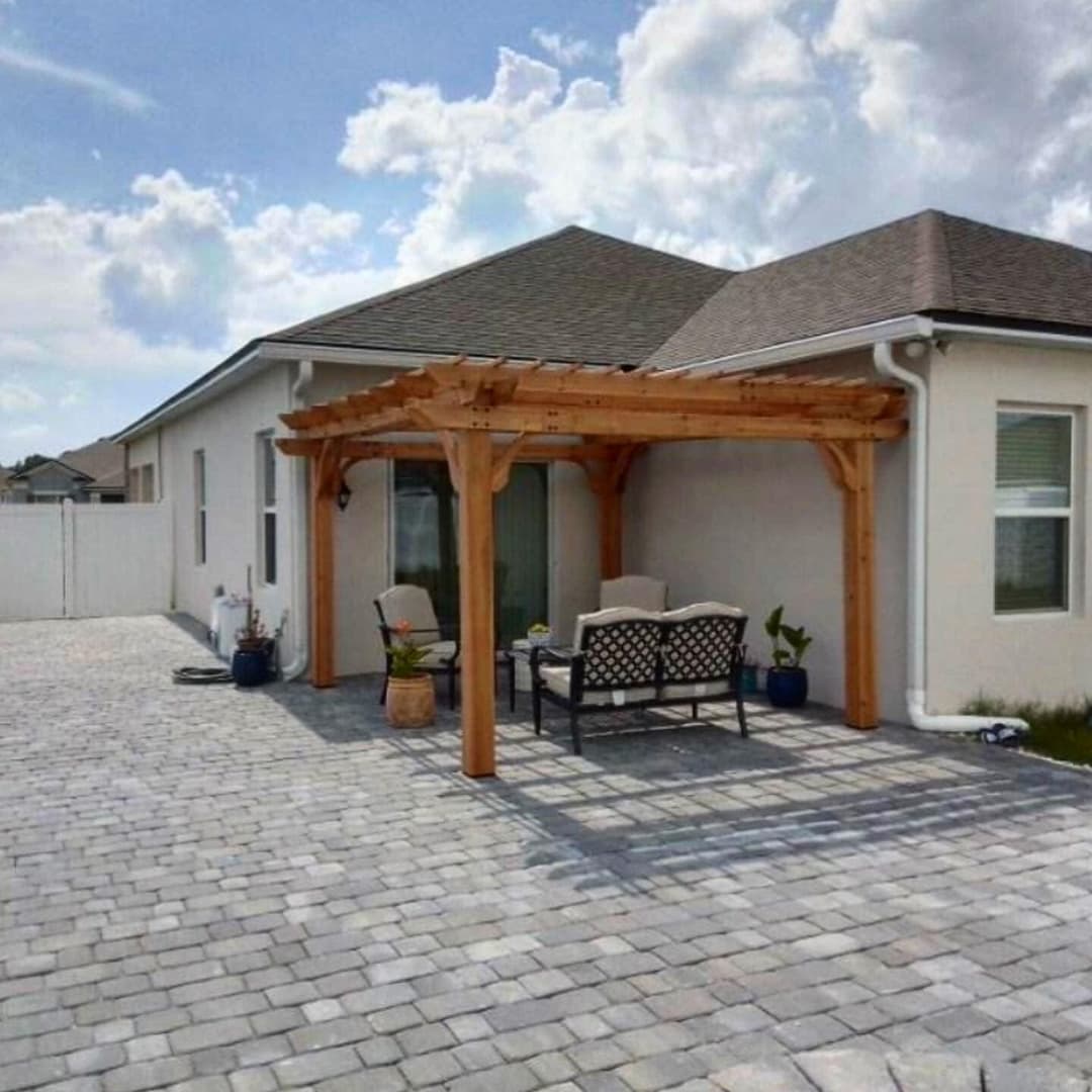 Wooden pergola with seating area and paver patio beside a modern house on a sunny day.