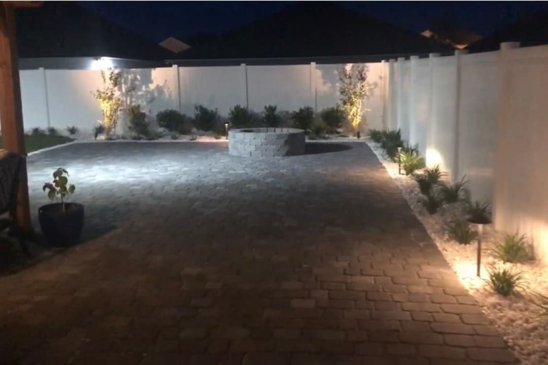 Paver patio with fire pit and landscape lighting in a modern backyard at night.