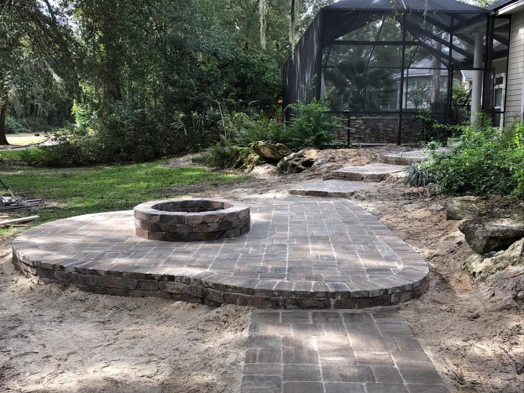Paved patio with circular fire pit surrounded by greenery and a screened patio in the background.