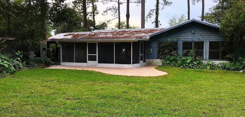 Blue house with screened porch, surrounded by greenery and trees in a spacious yard.