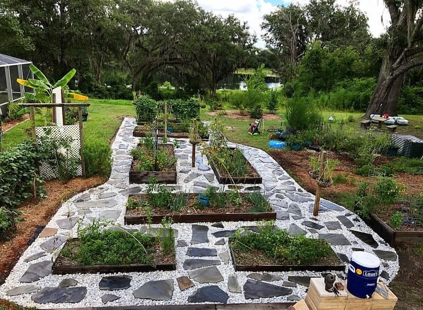 Lush garden layout with raised beds, pathways, and greenery near a serene water backdrop.
