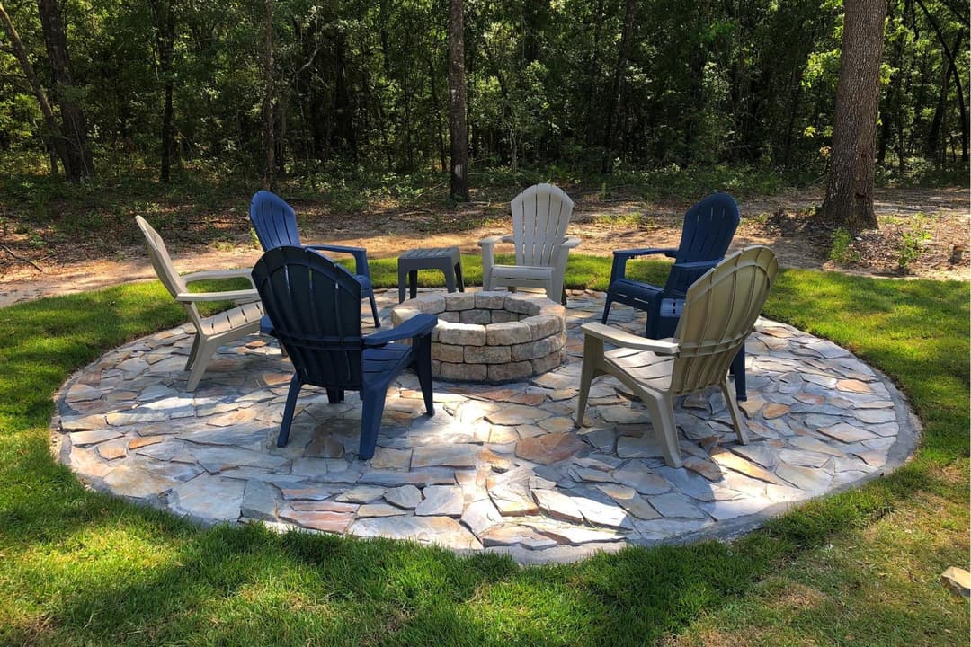Cozy outdoor fire pit area with stone seating and surrounding chairs in a wooded setting.