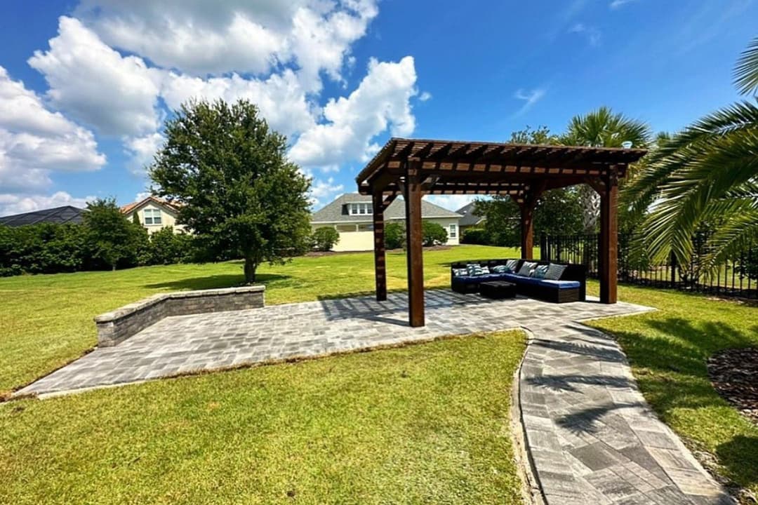 Outdoor patio with a wooden pergola, seating area, and landscaped lawn under a blue sky.
