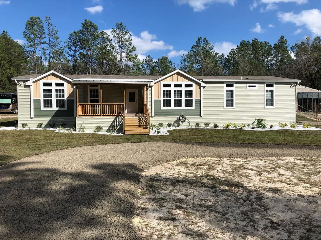 Modern manufactured home with a front porch, landscaped yard, and surrounding trees.