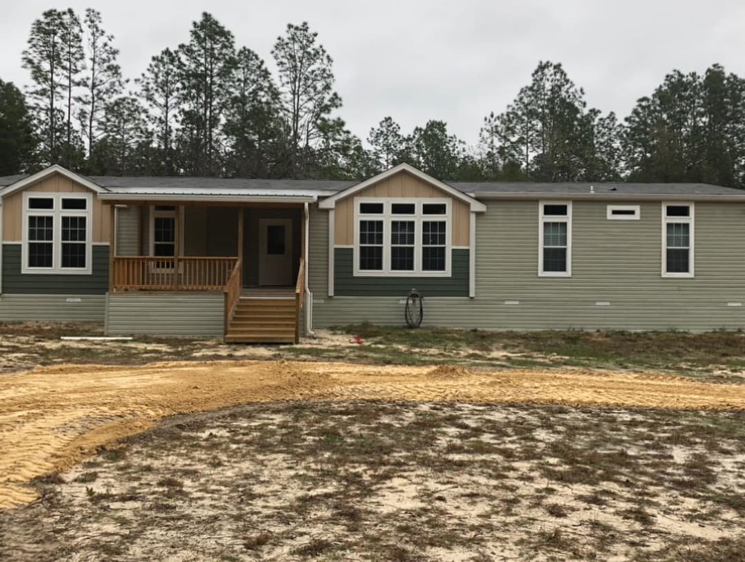 New manufactured home with a wooden porch, surrounded by trees and natural landscaping.