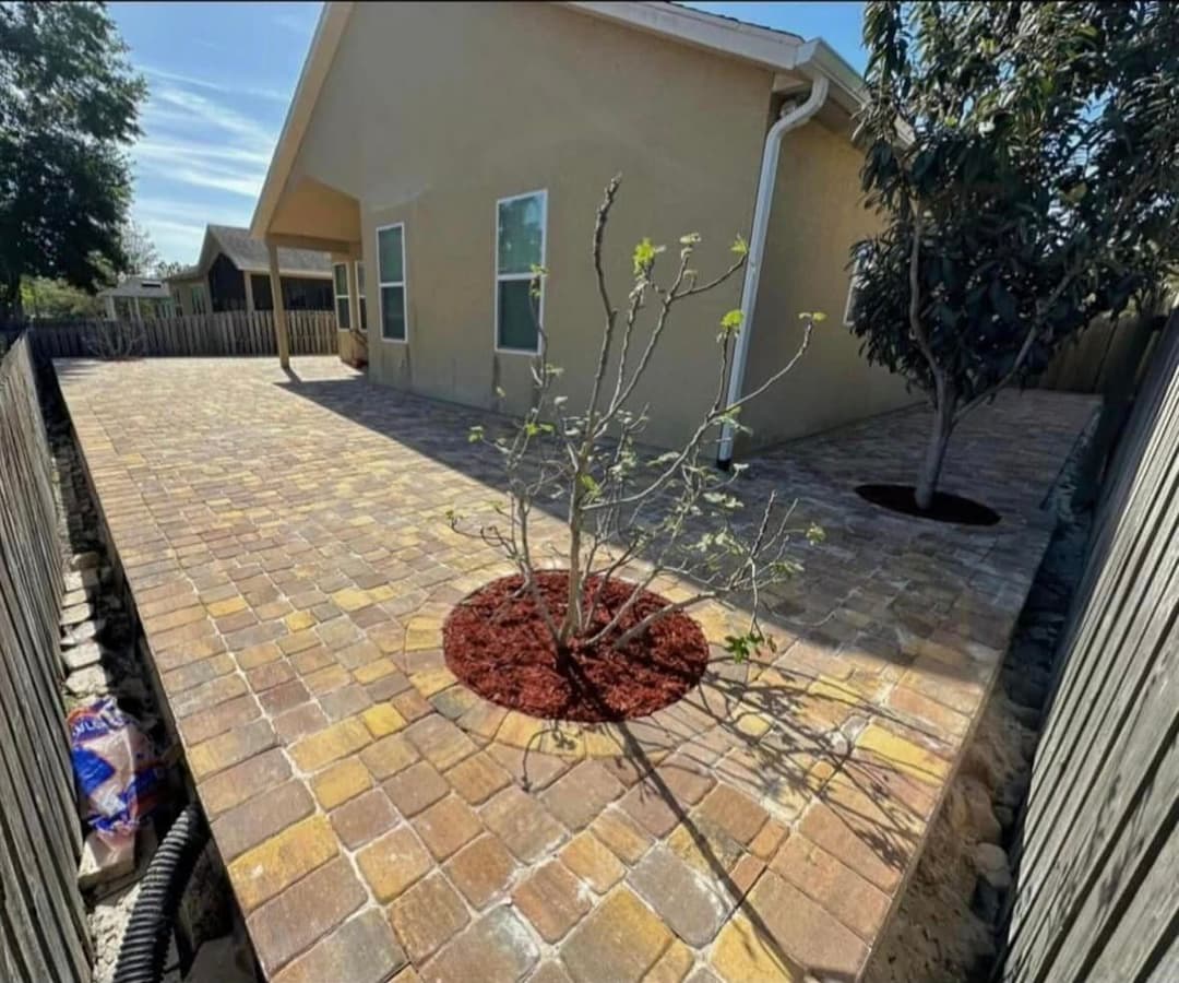 Patio with stone pavers, red mulch, and young tree near a house and wooden fence.