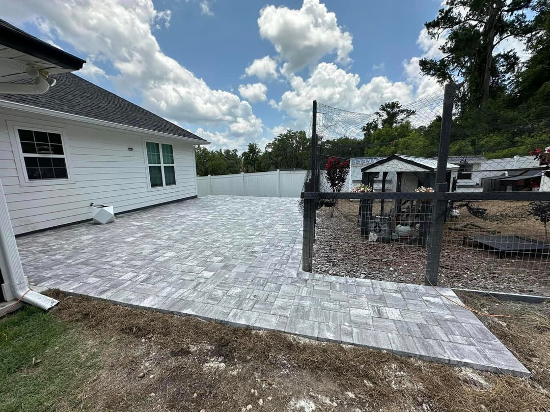 Paved patio area beside a fenced yard and coop under a blue sky with fluffy clouds.