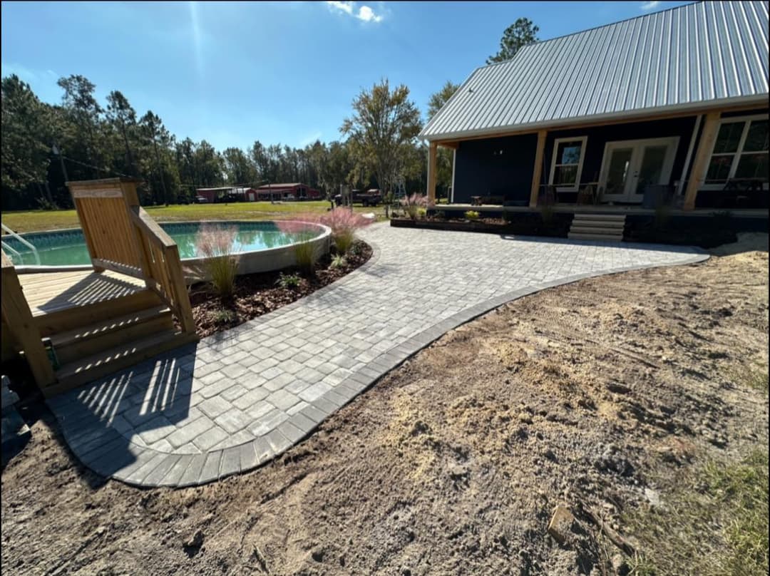Paved walkway curves around pool, leading to modern house with patio and landscaped garden.