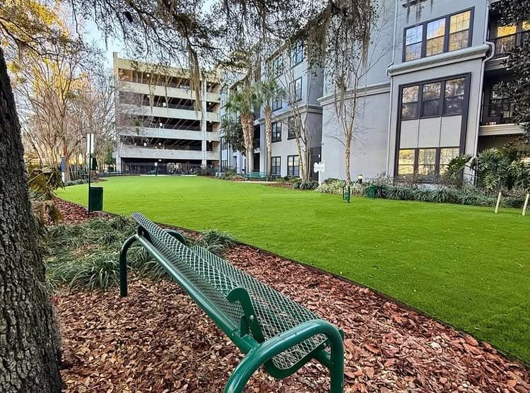 Green park bench on artificial turf with nearby parking garage and trees. Urban outdoor space.