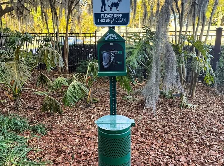 Dog waste station in a park with a sign urging cleanliness amidst leafy surroundings.