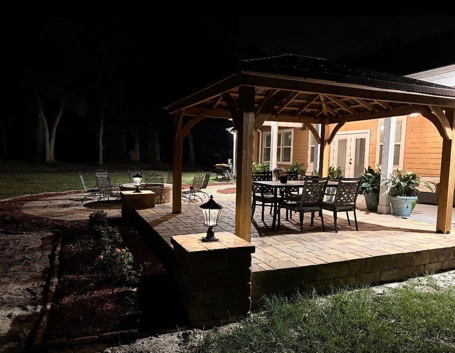 Illuminated outdoor patio with a pergola, seating area, and landscaped garden at night.