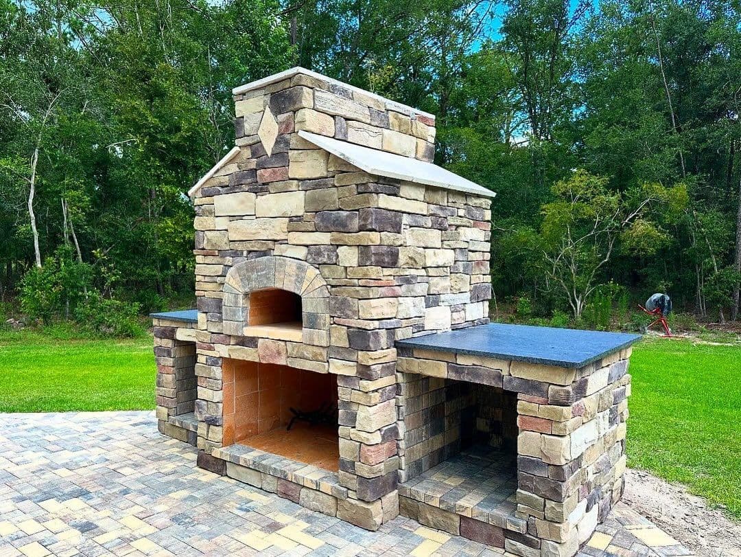 Outdoor stone pizza oven with chimney, surrounded by greenery and paving stones. Ideal for grilling.