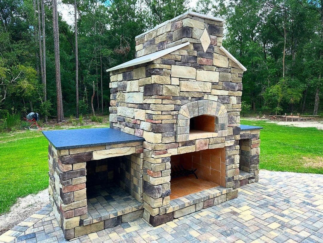 Outdoor stone pizza oven with chimney surrounded by lush greenery and paved patio.