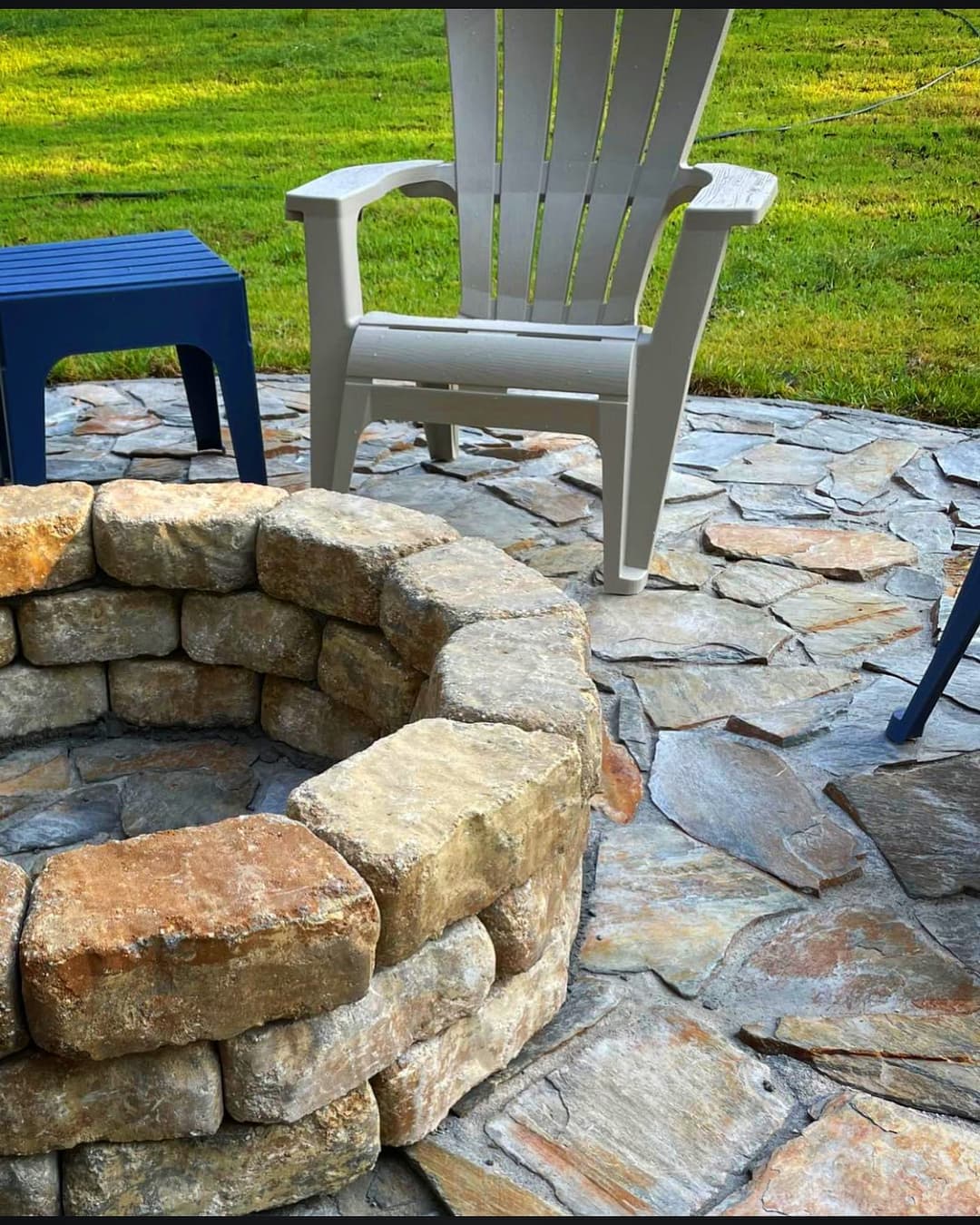 Stone fire pit surrounded by patio stones and outdoor chairs in a grassy yard.